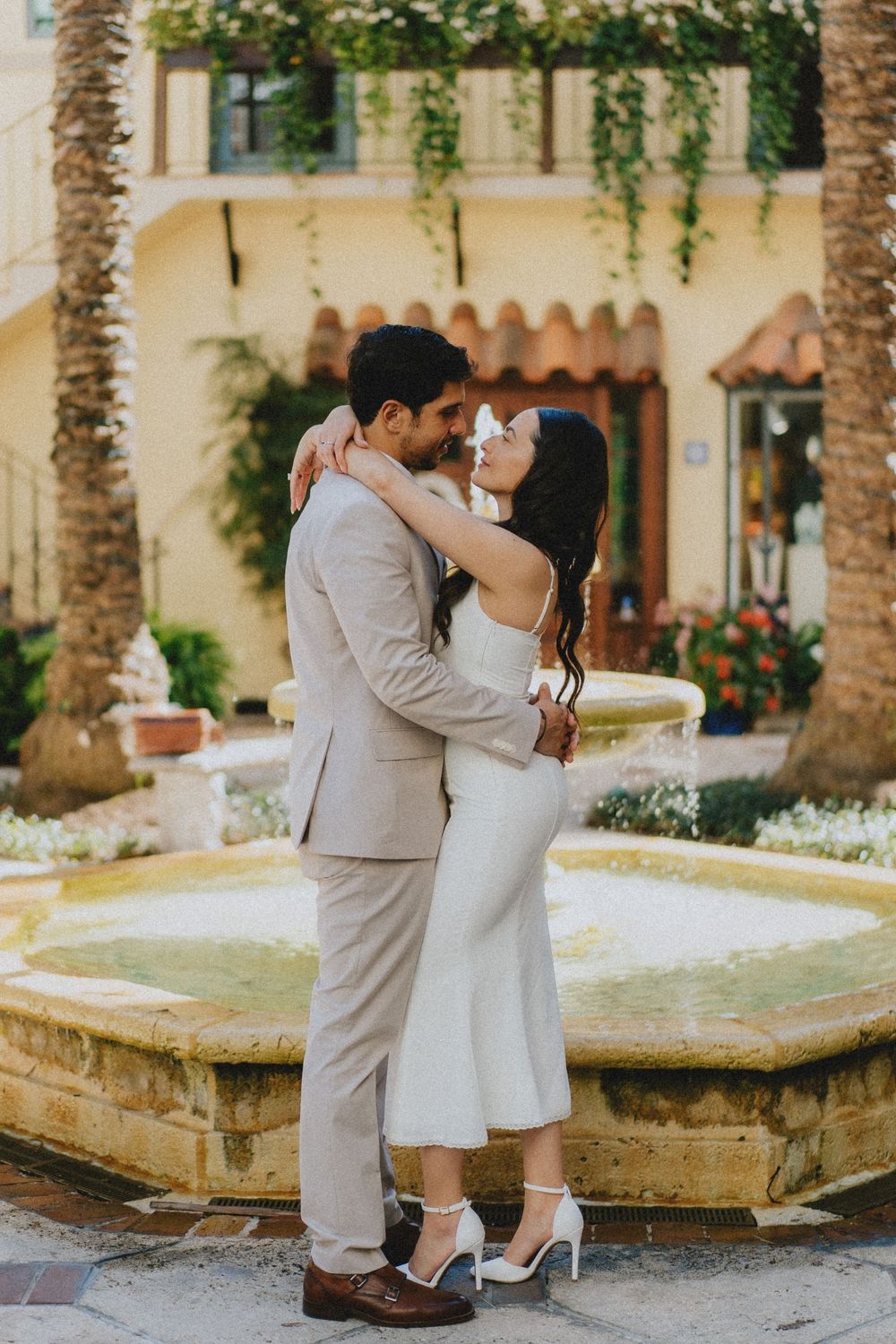 Embracing couple in formal wear share a tender moment by an ornate fountain in a Spanish-style courtyard.