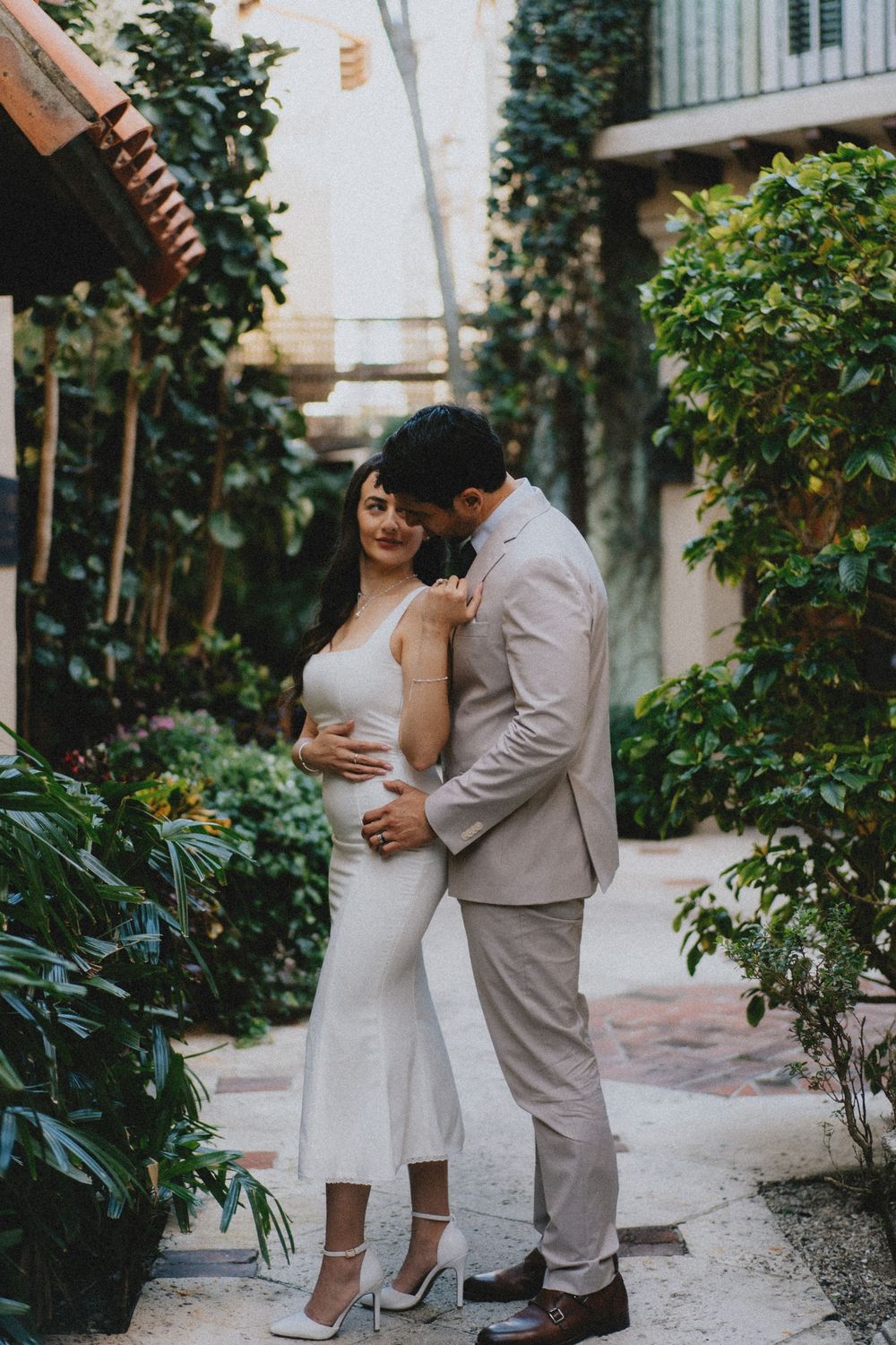 A romantic couple embraces on a garden path with lush greenery and white walls surrounding them.