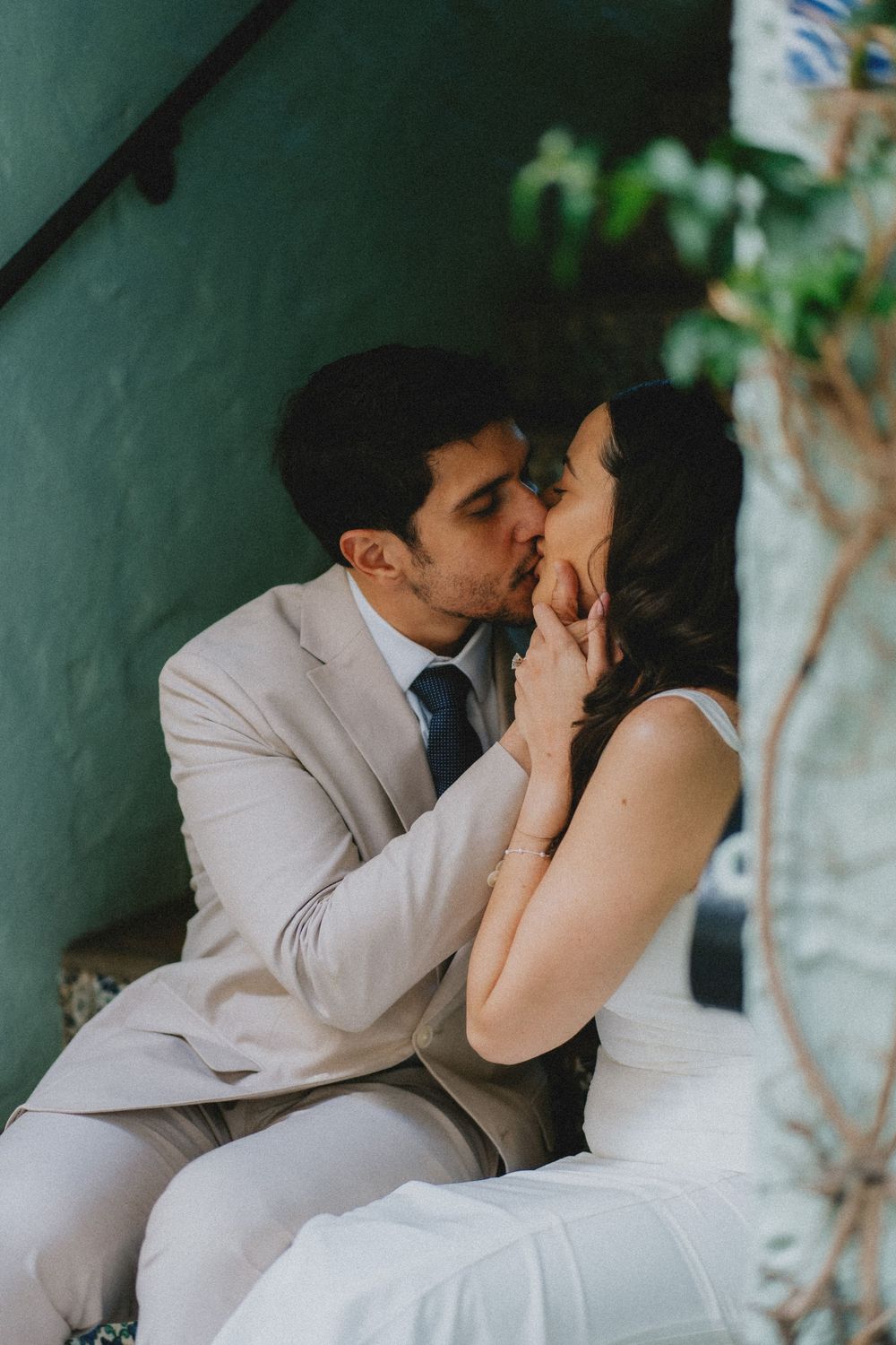 A romantic couple in formal attire share an intimate kiss against a green wall with plants in soft lighting.