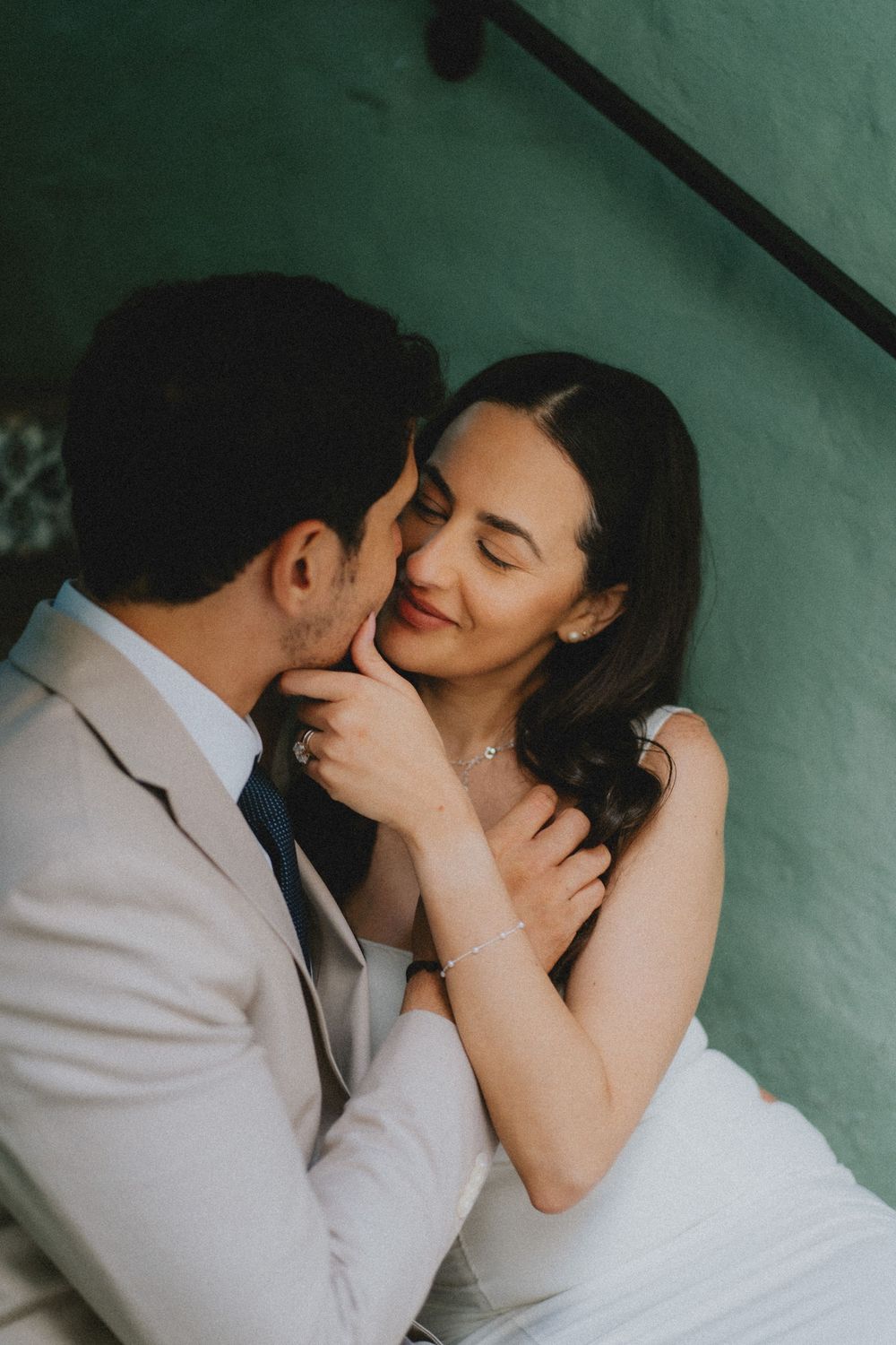 A tender romantic moment between newlyweds against a green wall, with the bride wearing a white dress and groom in beige suit.