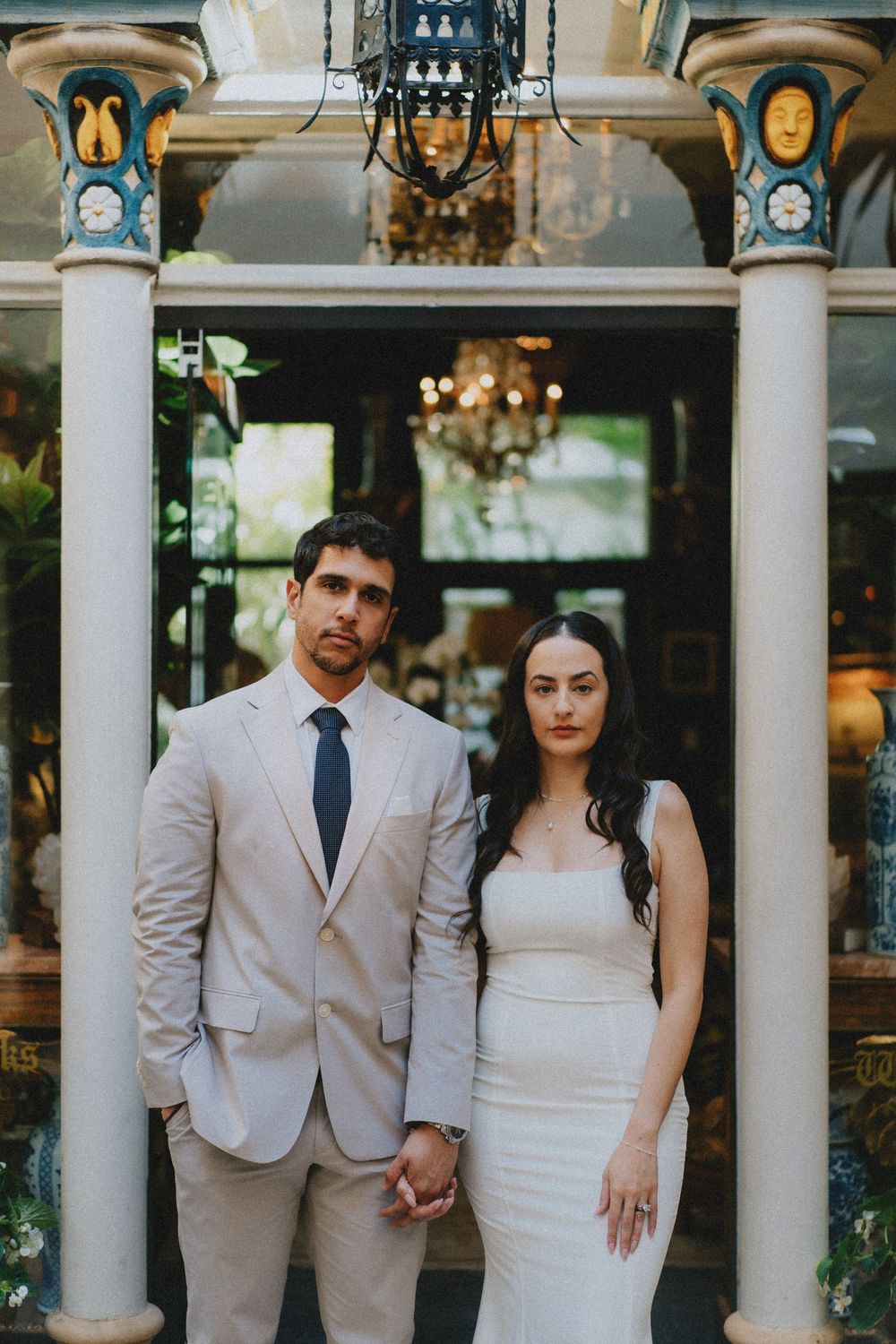A well-dressed couple poses beneath ornate columns and a chandelier in an elegant architectural setting.