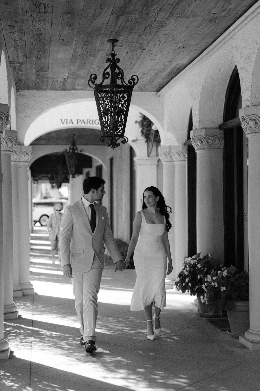 Couple walking hand in hand under arched colonnade with ornate wrought iron lantern and dappled sunlight casting shadows.