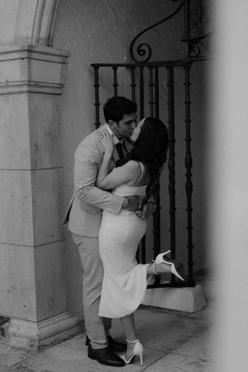 A couple in elegant attire share a romantic embrace against a stone wall with decorative iron railings.