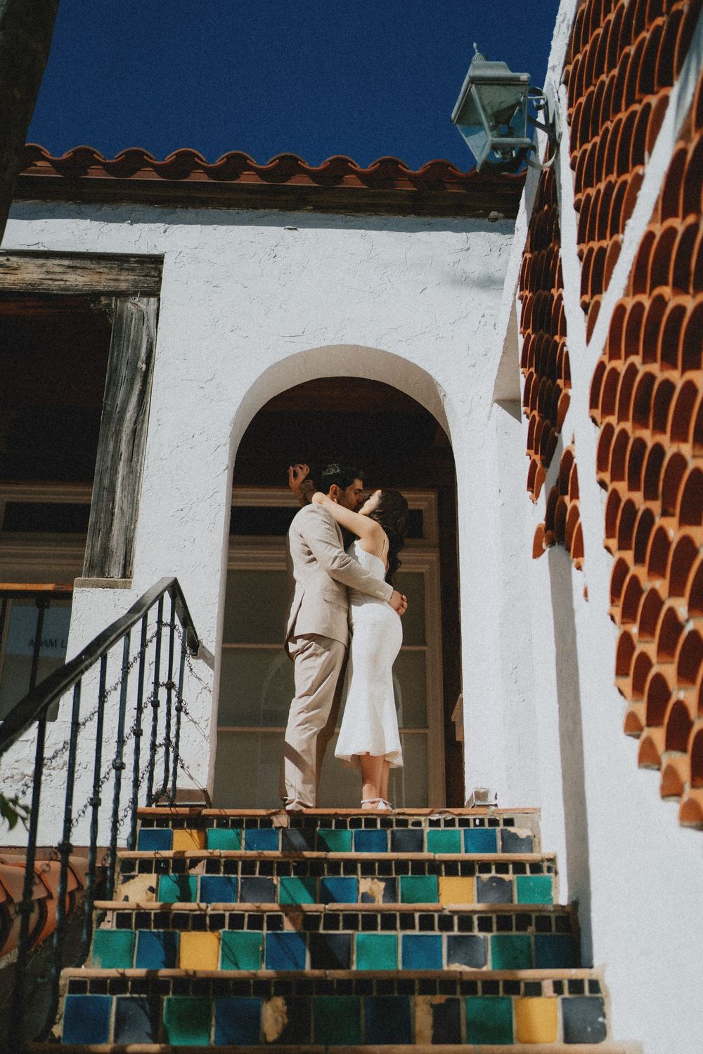 A couple embraces on colorful tiled steps in front of a Spanish colonial style building with white stucco walls.
