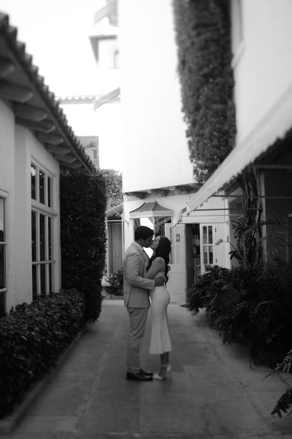 A vintage black and white photograph of a couple in an intimate moment along a narrow garden path between buildings.