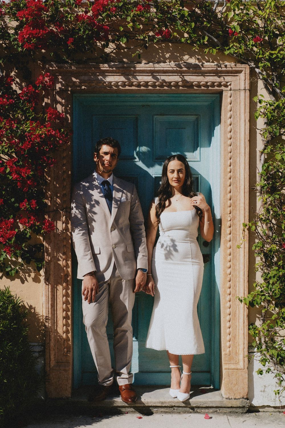 A couple stands in sunlight against a bright turquoise door framed by vibrant red bougainvillea flowers in an outdoor setting.