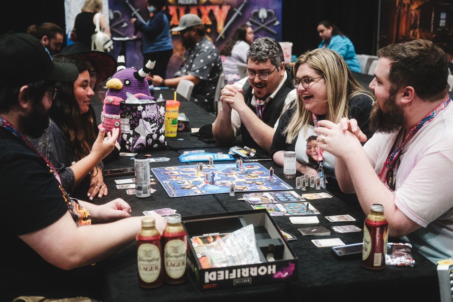 Group of people playing a board game at a table with drinks and cards in what appears to be a bar or gaming venue.