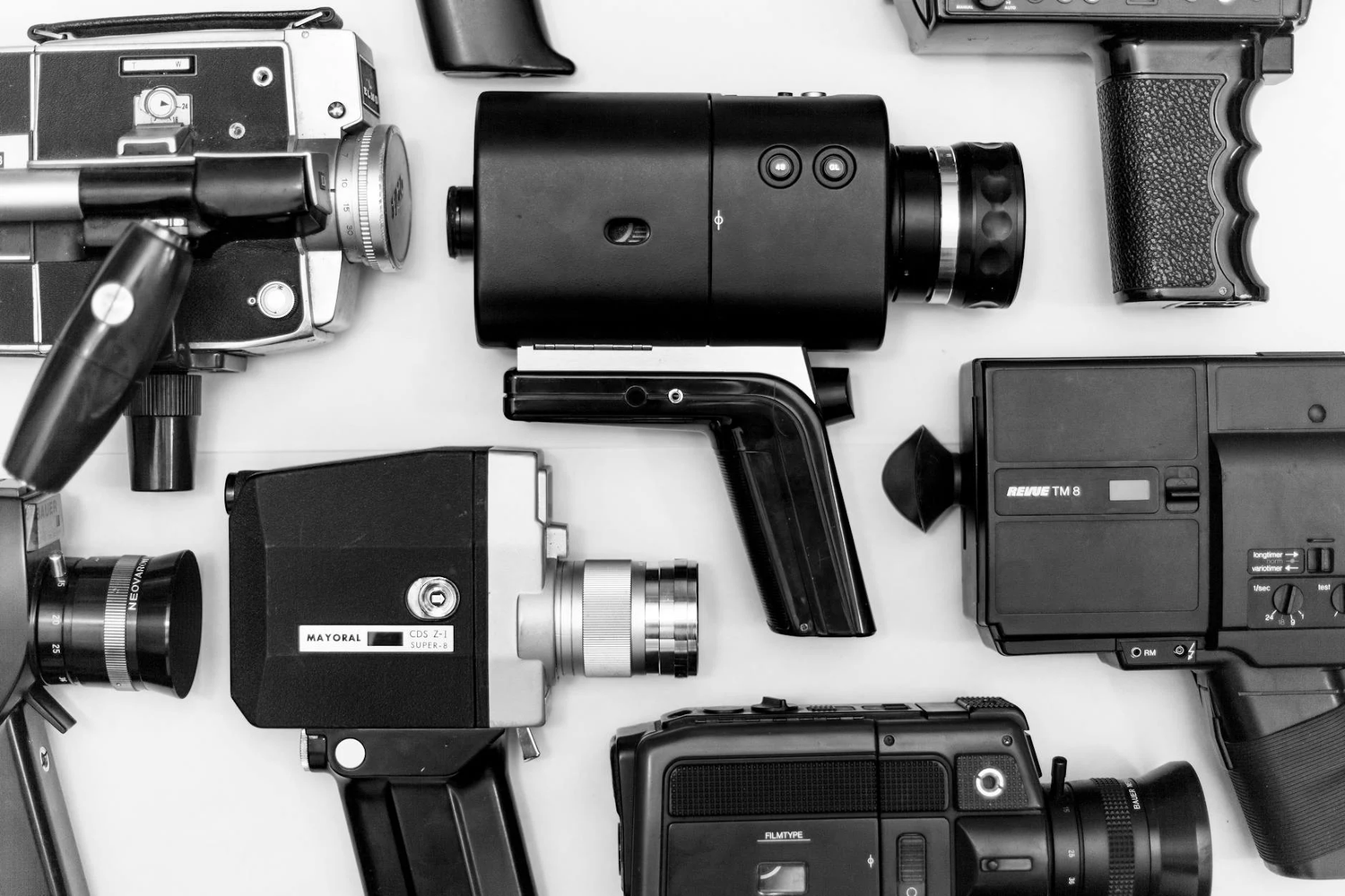 Black and white photo of vintage video cameras and recording equipment arranged in a flat lay composition on white background.