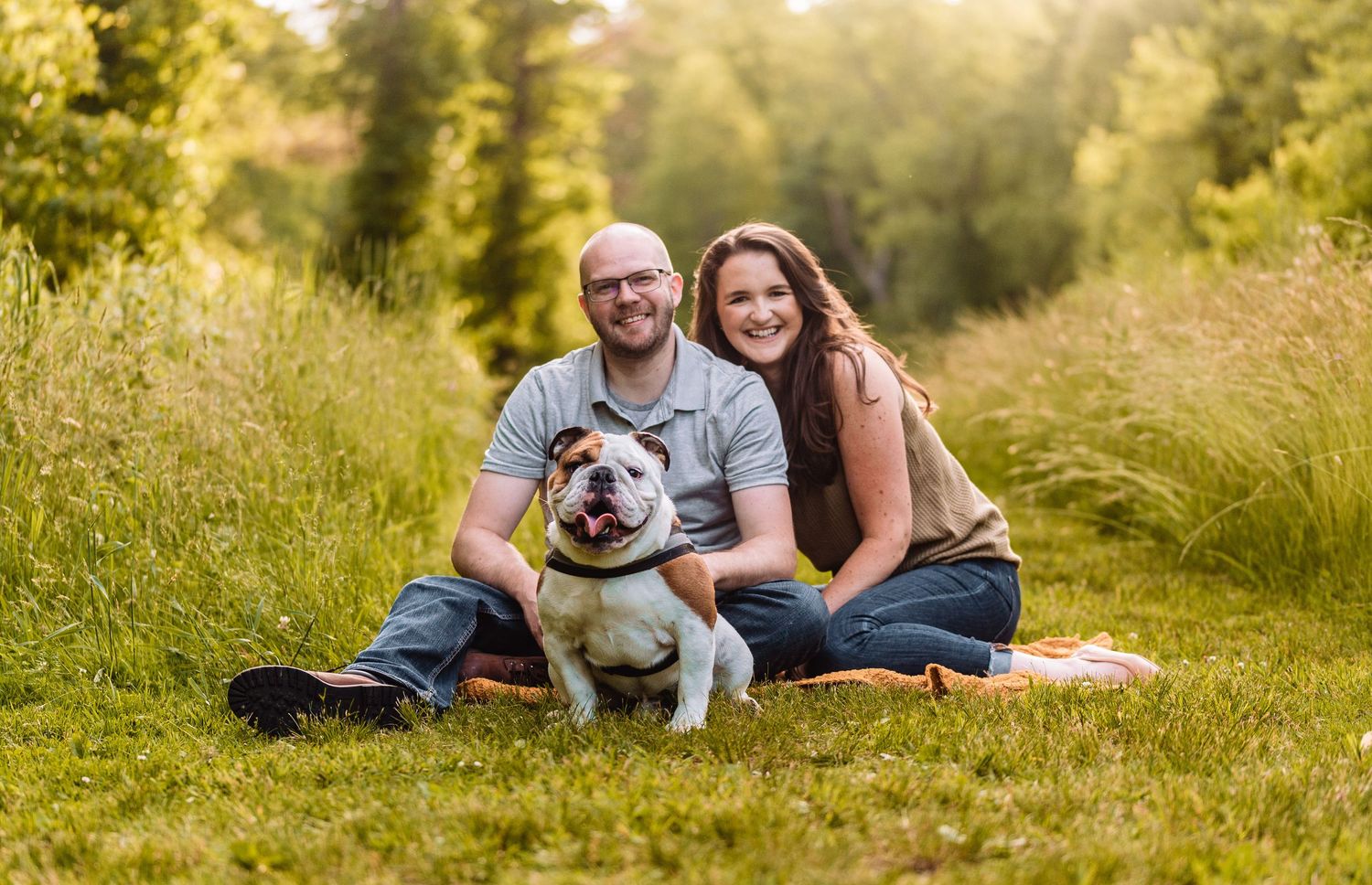 A couple sits in a grassy meadow during golden hour with their English bulldog on a blanket.