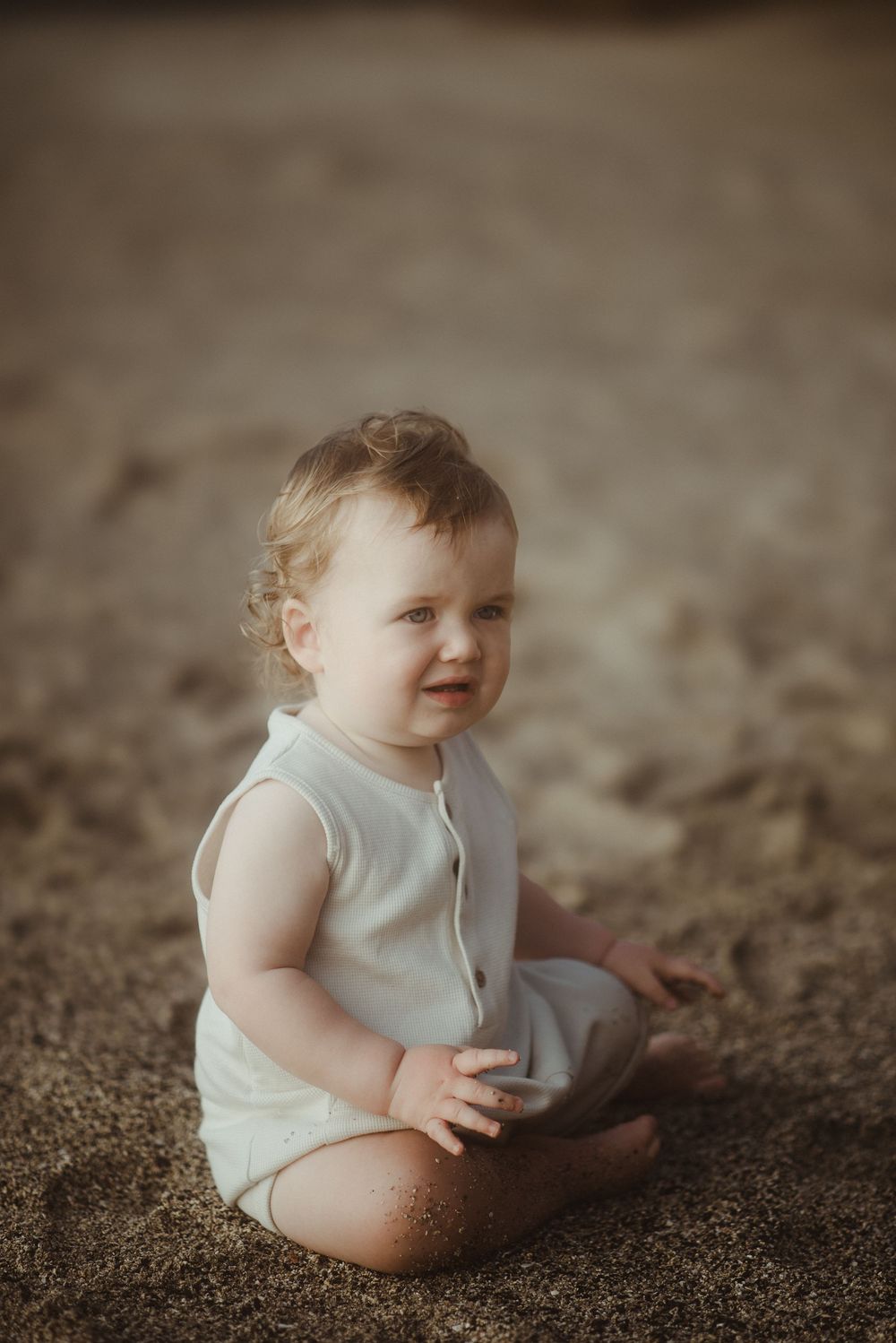 A small child wearing a light-colored romper sits contentedly on a brown sandy surface in warm lighting.