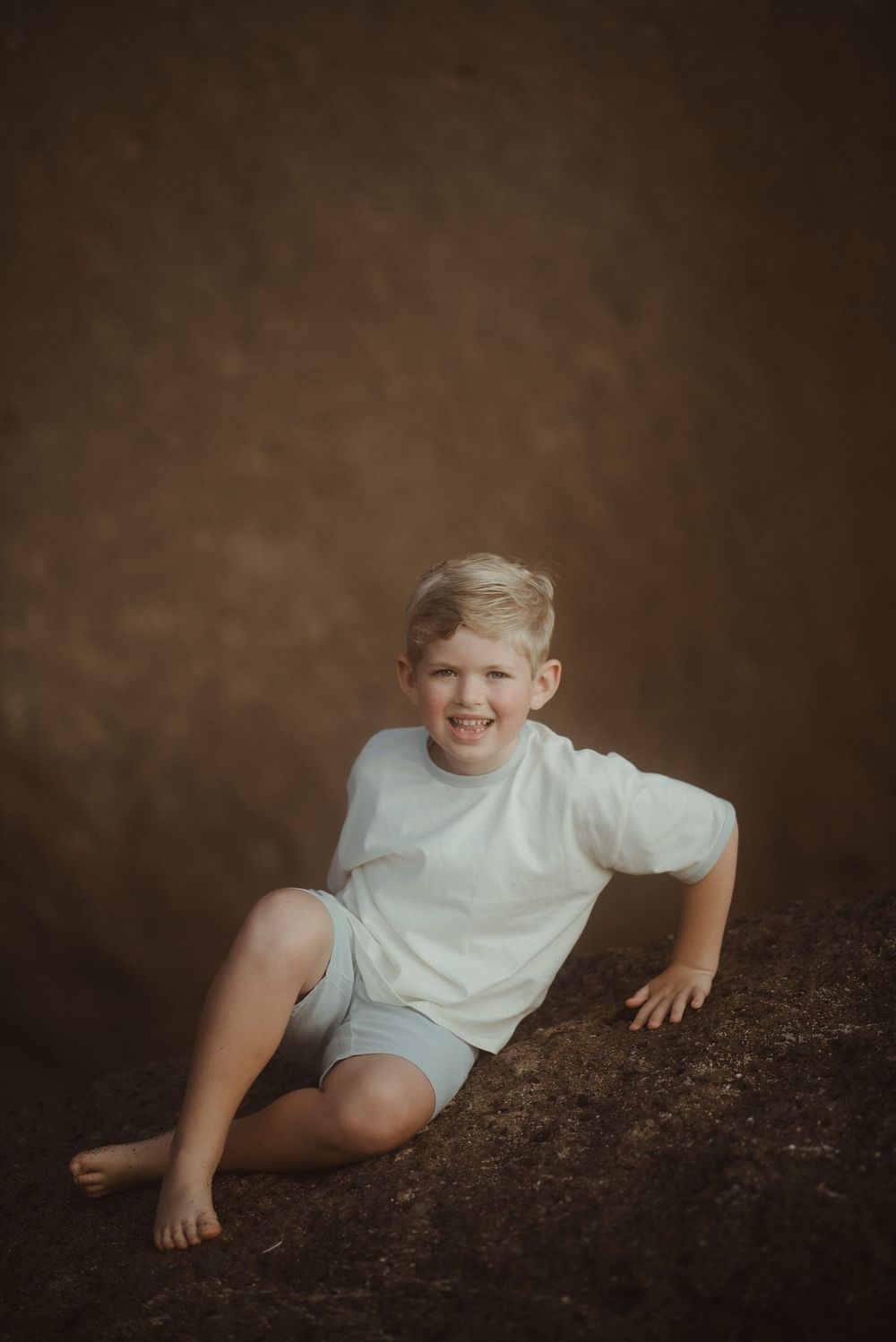 A young child in white clothing sits casually on a dark textured backdrop for a moody portrait.