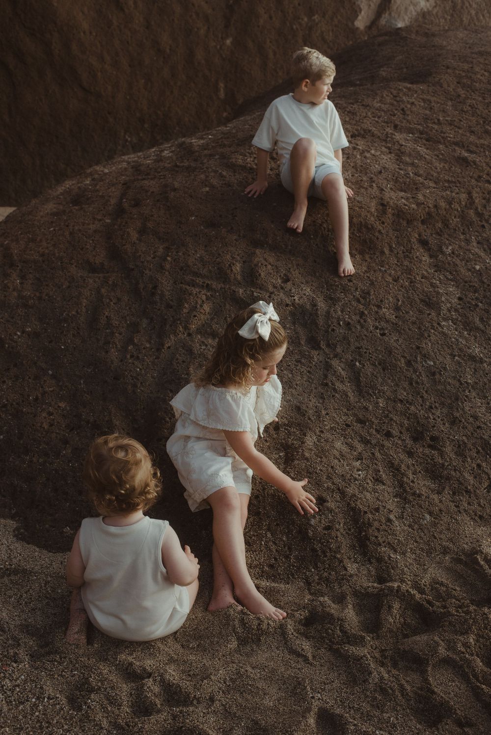 Three children in white clothing sit on rocky terrain during an outdoor photo session.