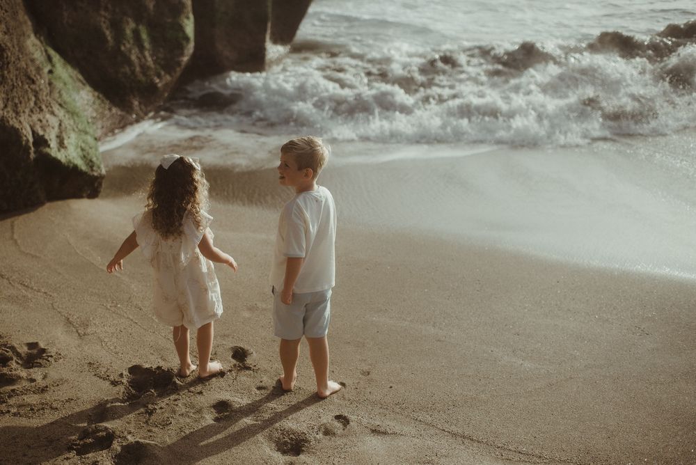 Two children stand at the edge of a sandy beach as waves crash against nearby rocks on an overcast day.