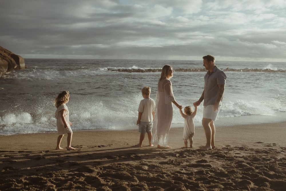 A family silhouetted against a dramatic cloudy beach scene stands together on dark sand as waves crash nearby.