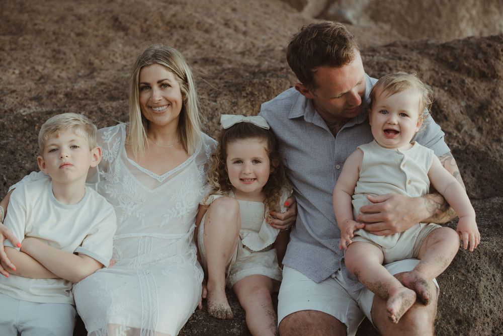 A loving family sits together on rocky terrain during a sunset photo session at the beach.