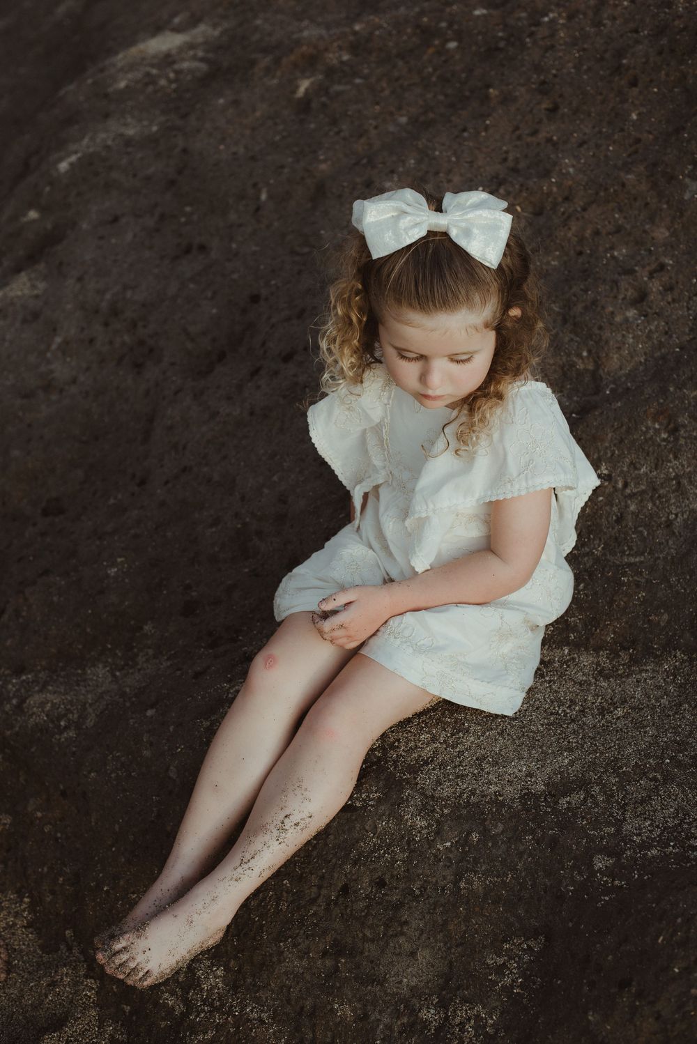 Artistic portrait of a child in white dress and bow sitting on dark ground with head bowed down in contemplation.