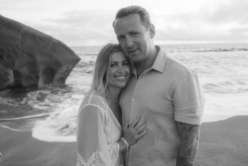 A black and white romantic portrait shows a couple embracing on a beach with cliffs visible in the background.