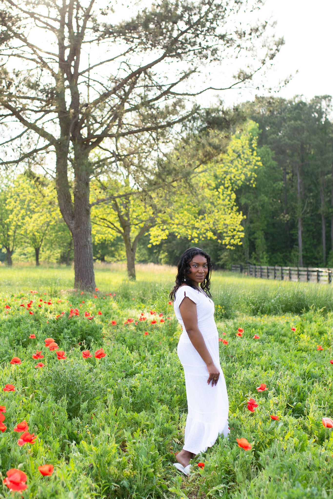 a Woman in a flowing white dress stands in a vibrant spring meadow filled with blooming red poppies and tall trees.