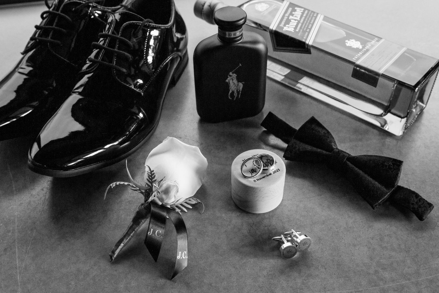 Black and white still life of formal dress shoe, cologne bottle, boutonniere, and accessories arranged on dark surface.