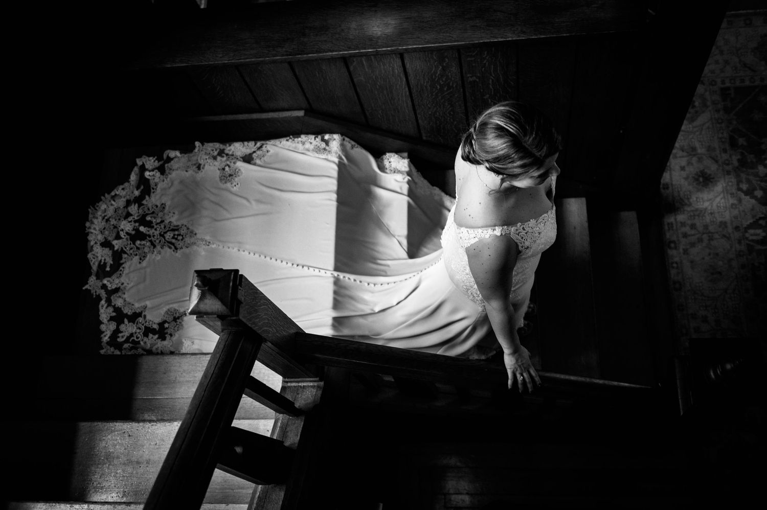 Black and white photograph of figure in flowing white dress descending dark wooden staircase in dramatic lighting.