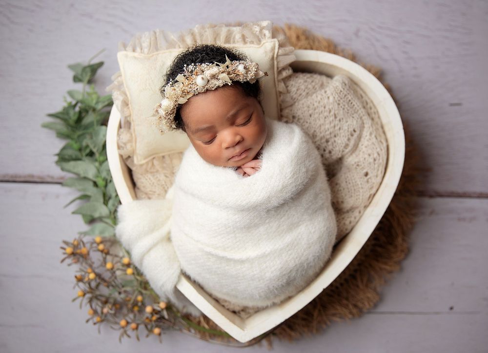 A sleeping newborn wrapped in white swaddling lies in a heart-shaped basket with floral decor and a delicate headpiece. During a photoshoot