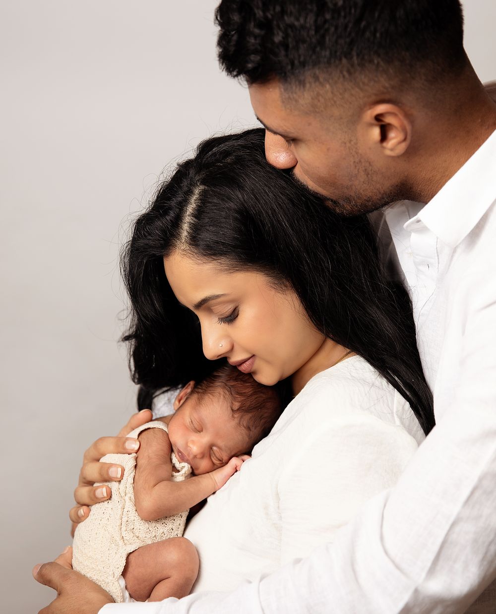Parents in white outfits lovingly embrace their sleeping newborn baby in a tender family portrait against a light background.