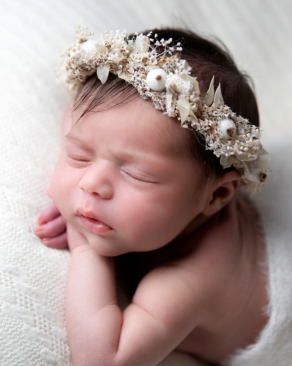 Sleeping newborn wearing delicate white floral and pearl crown headpiece on soft white fabric. During a photoshoot in Sidcup