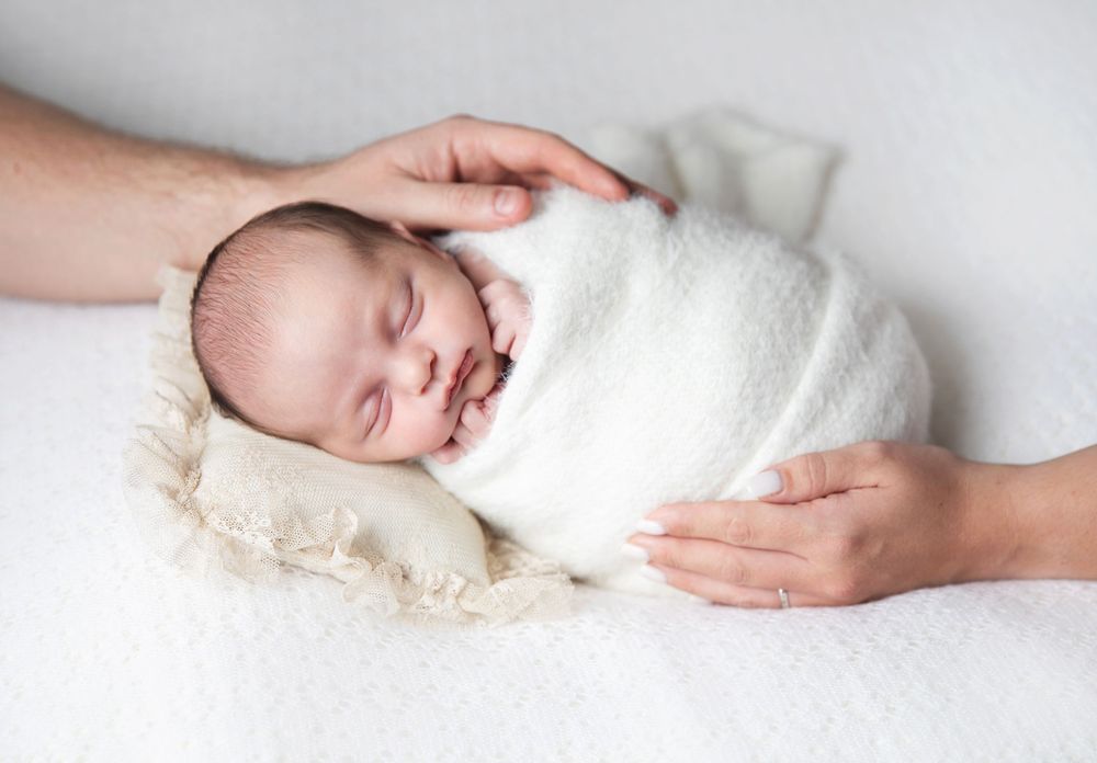A newborn sleeping peacefully on a white pillow, wrapped in a soft white blanket with gentle hands supporting. During a photoshoot in Sidcup