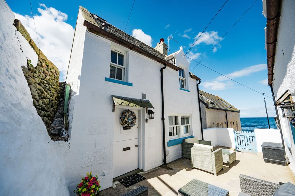 Charming white coastal cottage with blue trim and a wreath on the door overlooking the sea on a sunny day.