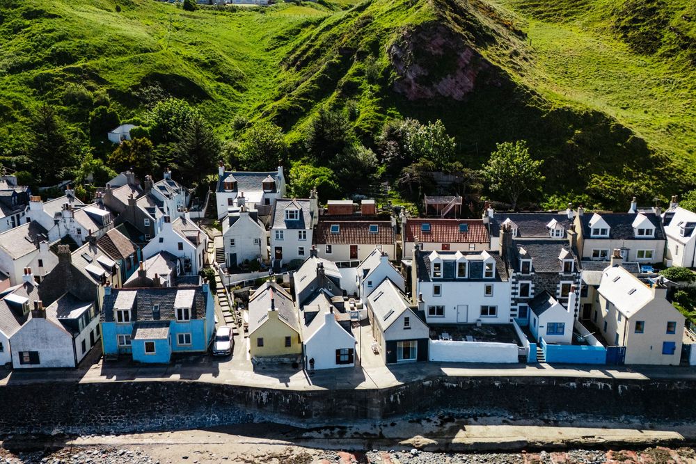 Aerial view of white seaside cottages lined along curved harbor wall with green hills above.