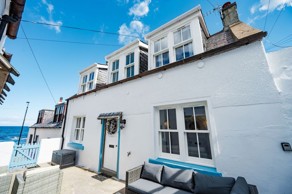 White coastal cottage with blue trim and outdoor seating area against bright sky.