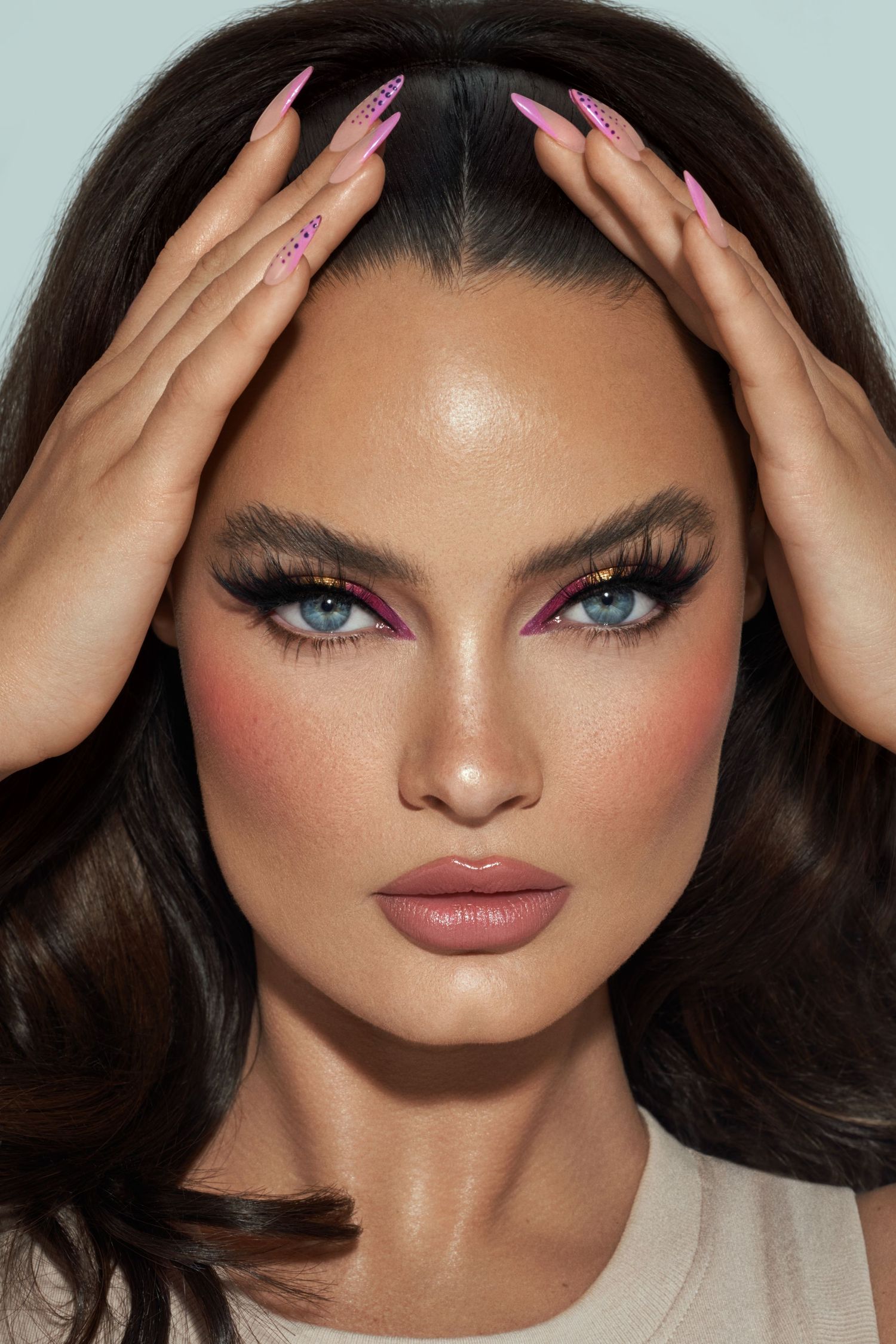 Glamour beauty shot with pink nails, dramatic eye makeup and voluminous hair against light background.