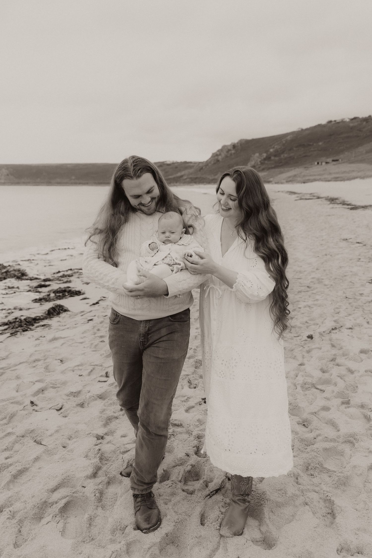 A person in a white sweater embraces and nuzzles a bundled baby while standing outdoors in soft lighting.