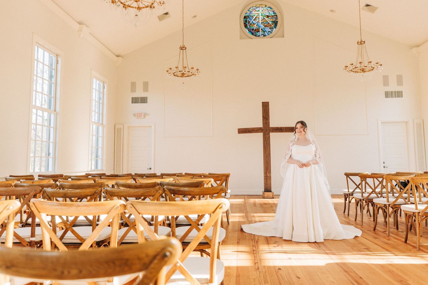 A sunlit chapel interior with wooden cross-back chairs and a wooden cross mounted on the white wall.