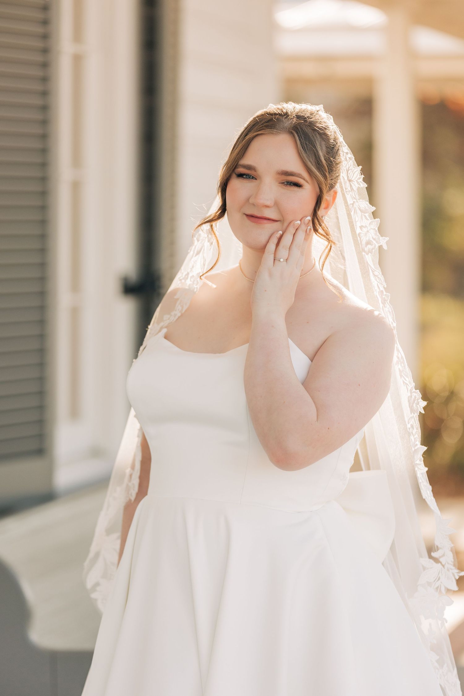 A person in a white wedding dress and veil stands in soft sunlight against a building exterior.