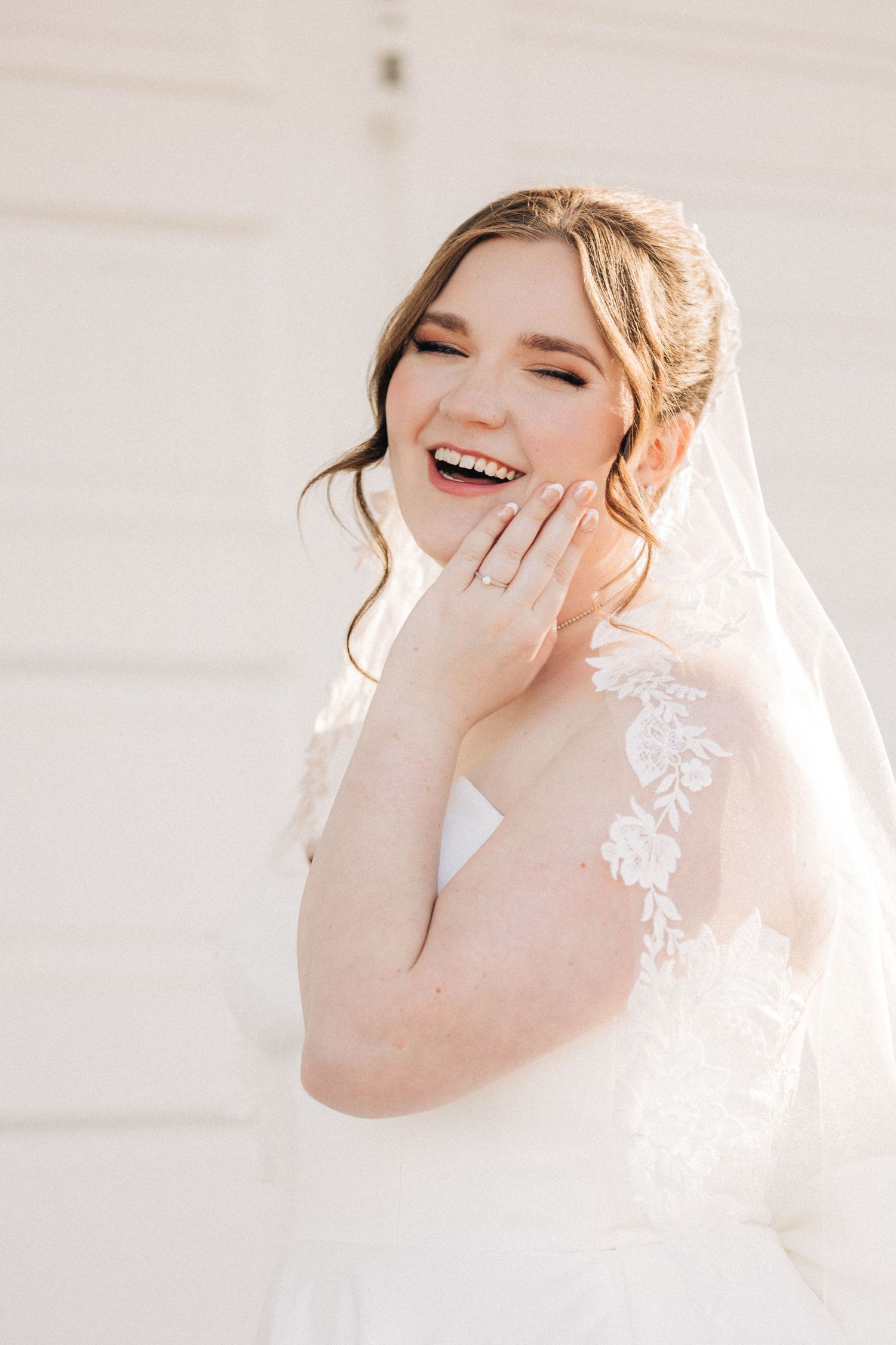 A joyful bride in a white lace wedding dress and veil laughs with genuine emotion against a light background.