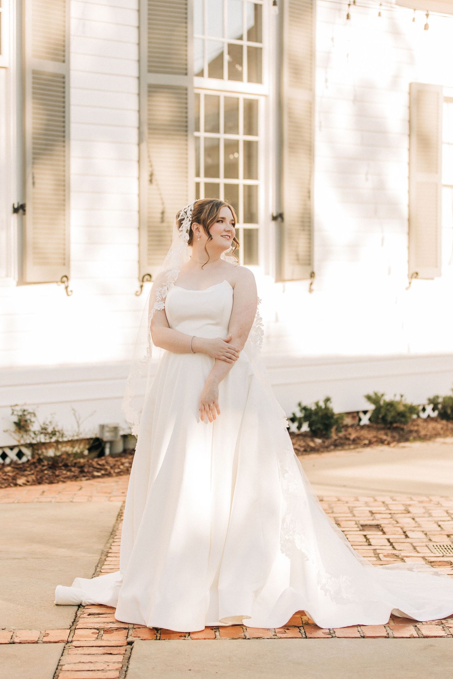 Ethereal bridal portrait with off-shoulder white gown standing on brick pathway by white building with shutters.