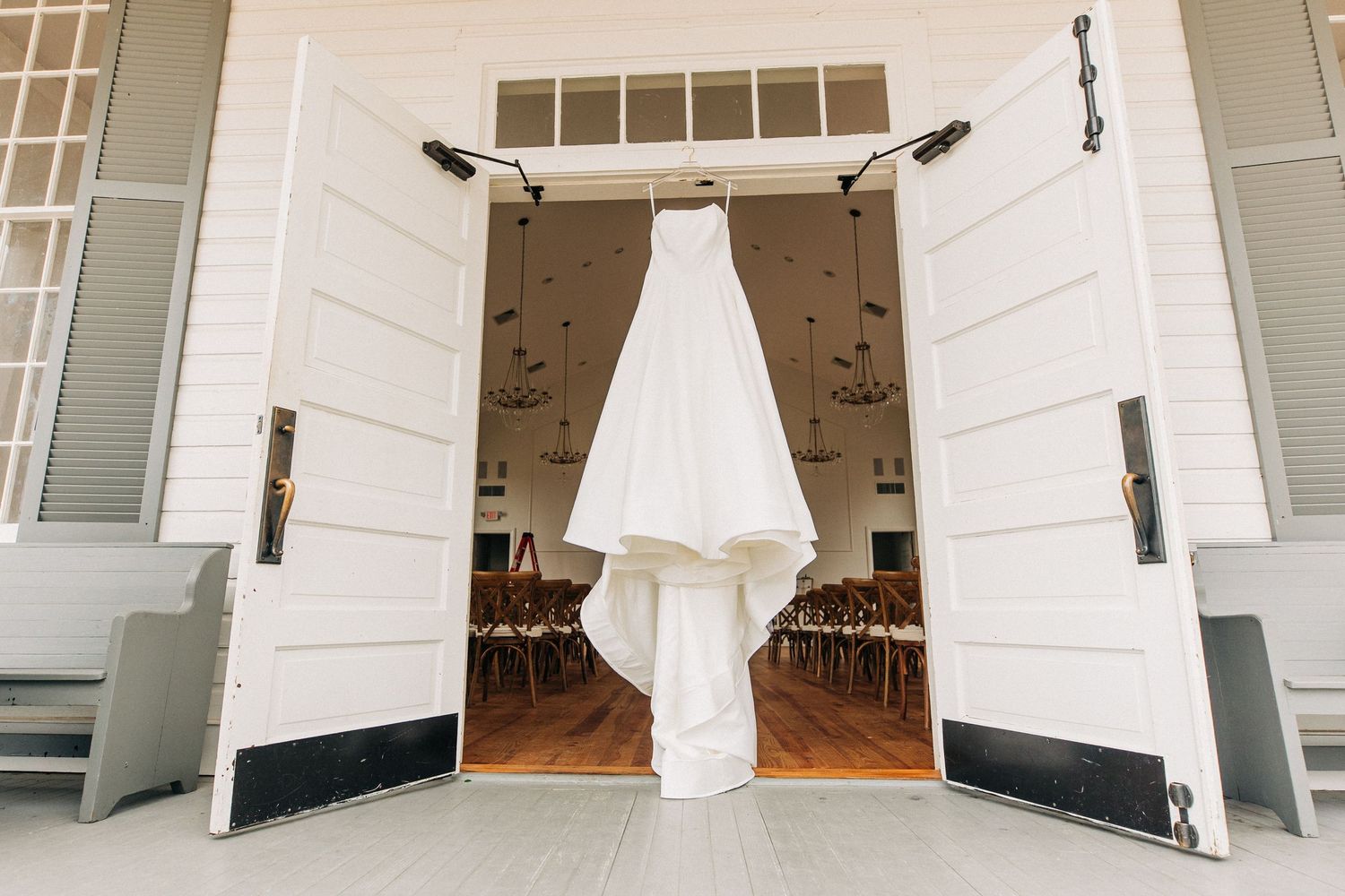 An elegant white wedding dress hangs from rustic white barn doors at a venue entrance.