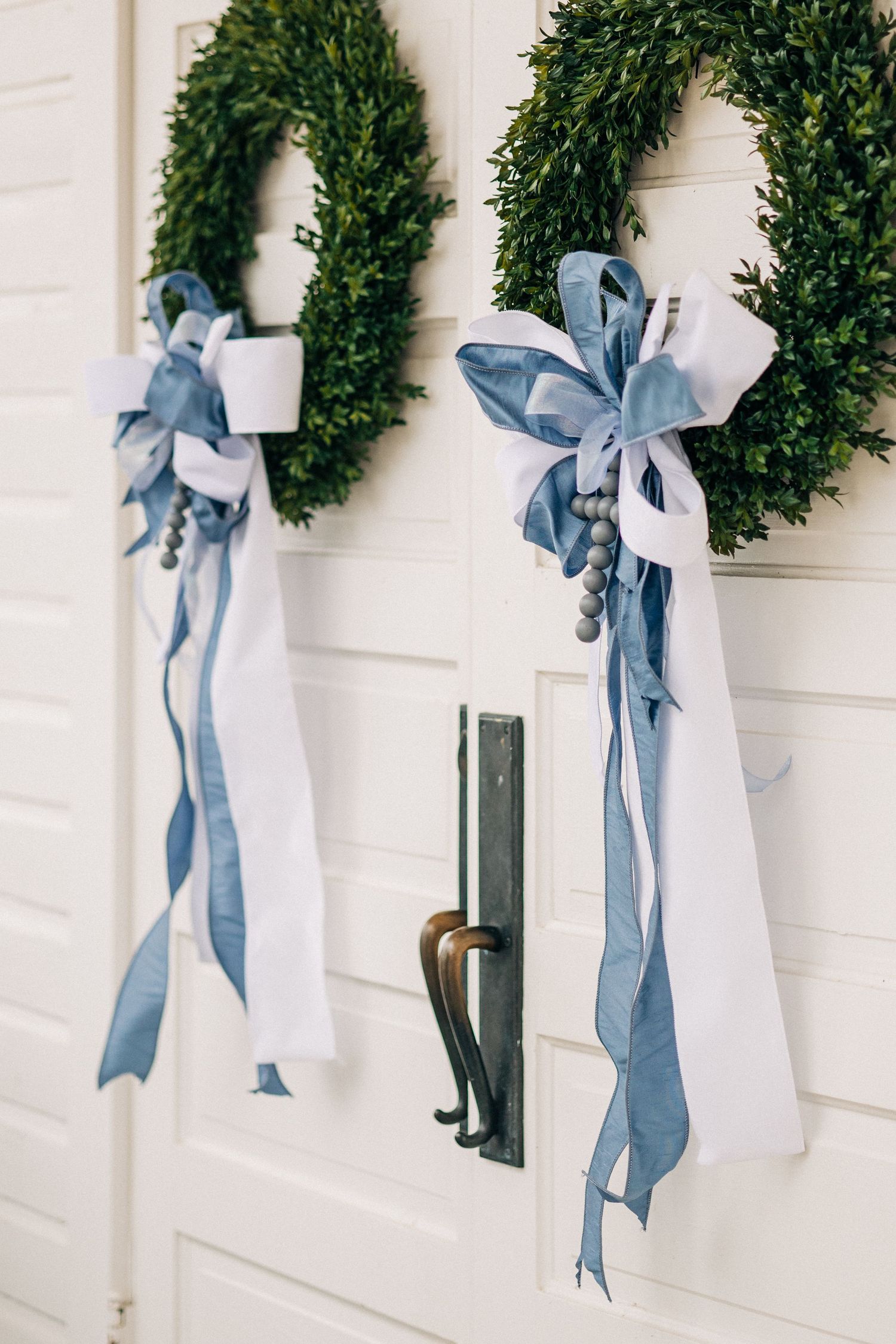 Two elegant boxwood wreaths decorated with white fabric and dusty blue ribbons hanging on a white door with black hardware.