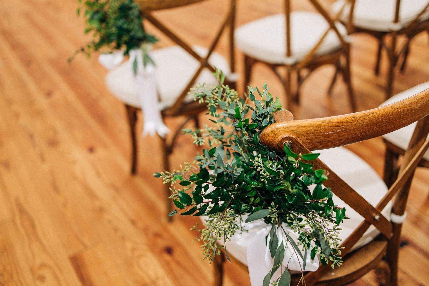 Cross-back wooden chair decorated with fresh greenery and white fabric for a rustic wedding ceremony setup.
