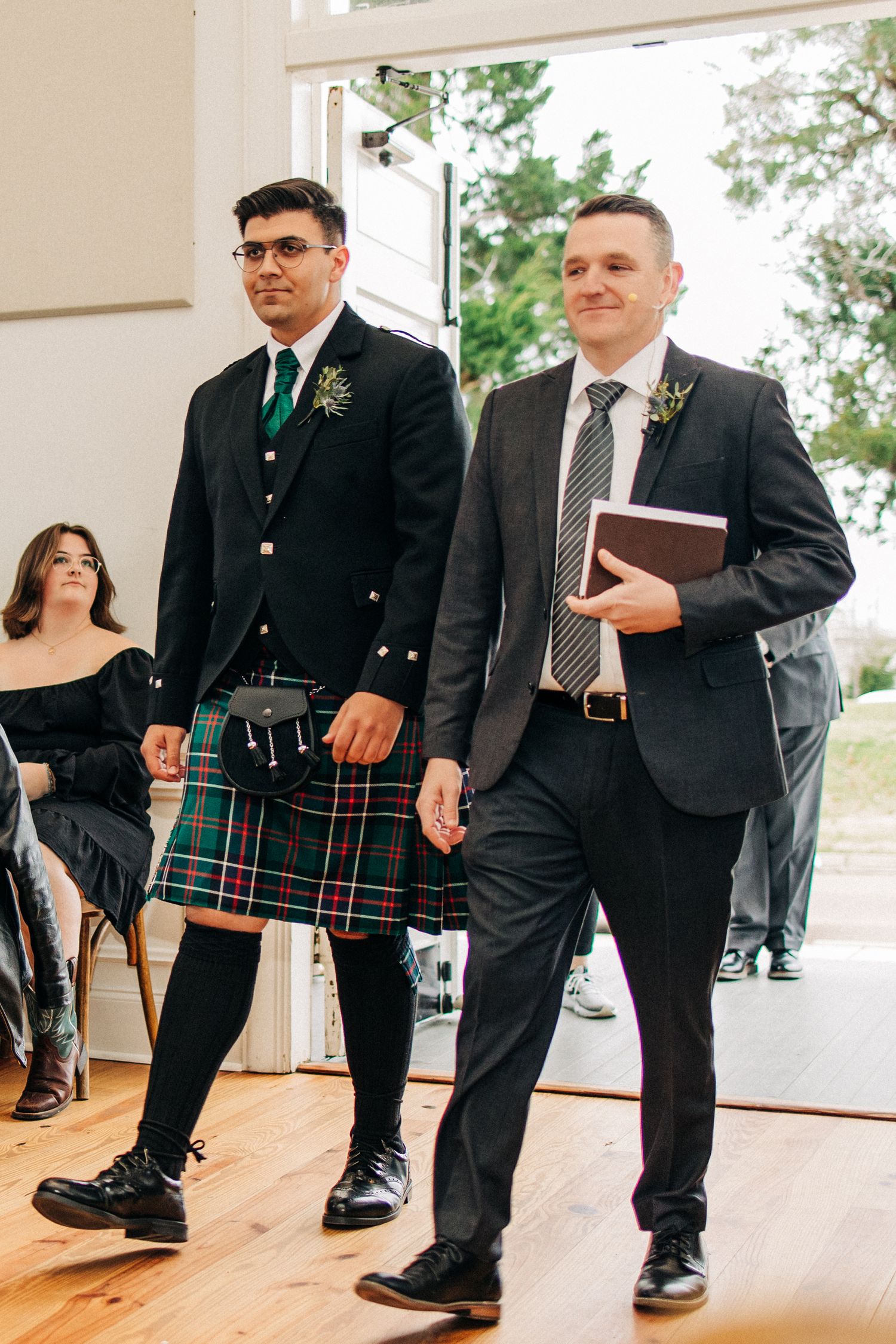 Two people walking down an aisle at a wedding ceremony, one wearing a traditional kilt and another in a dark suit.