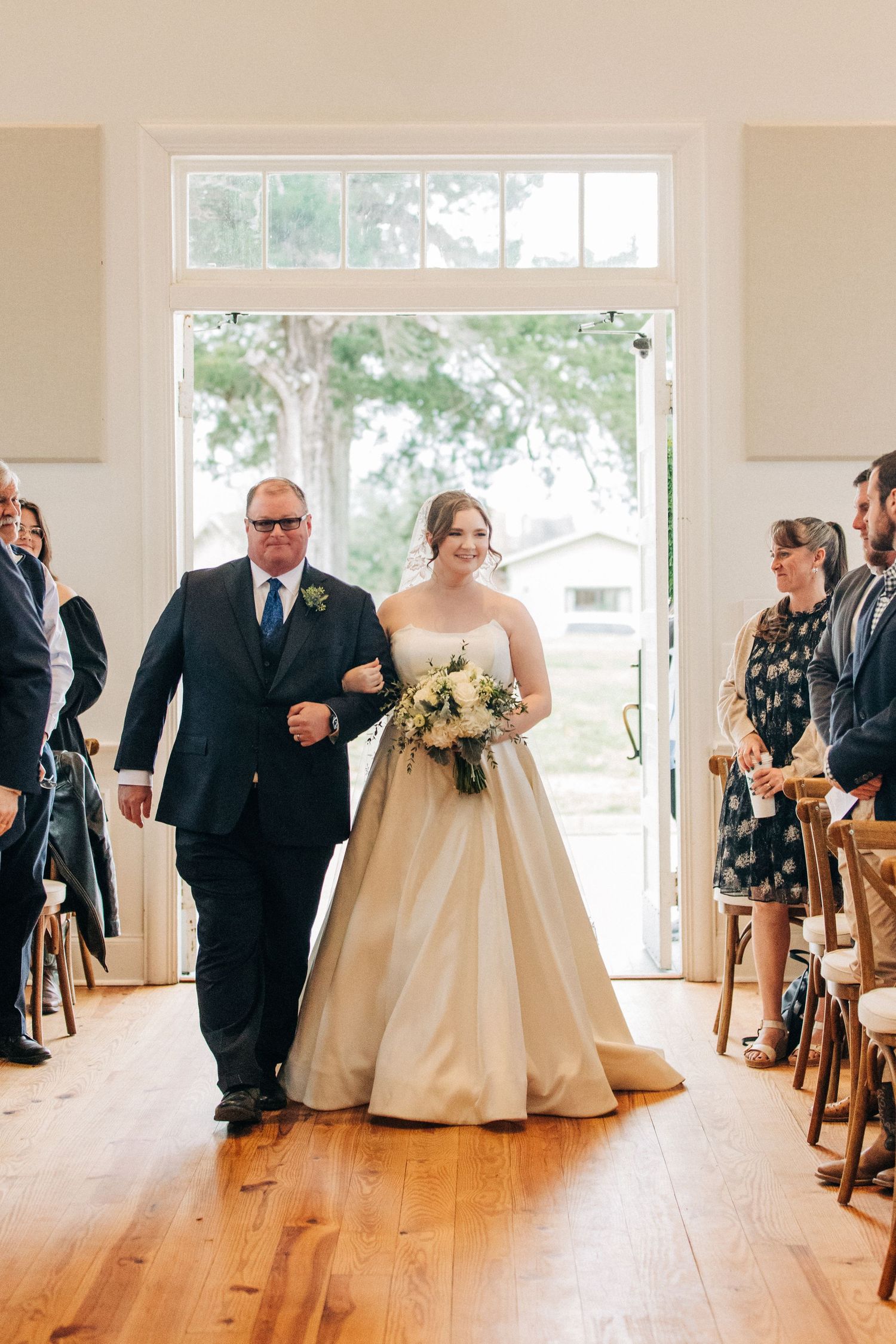 A couple walking down a wooden floor aisle surrounded by guests during a wedding ceremony in a bright indoor venue.