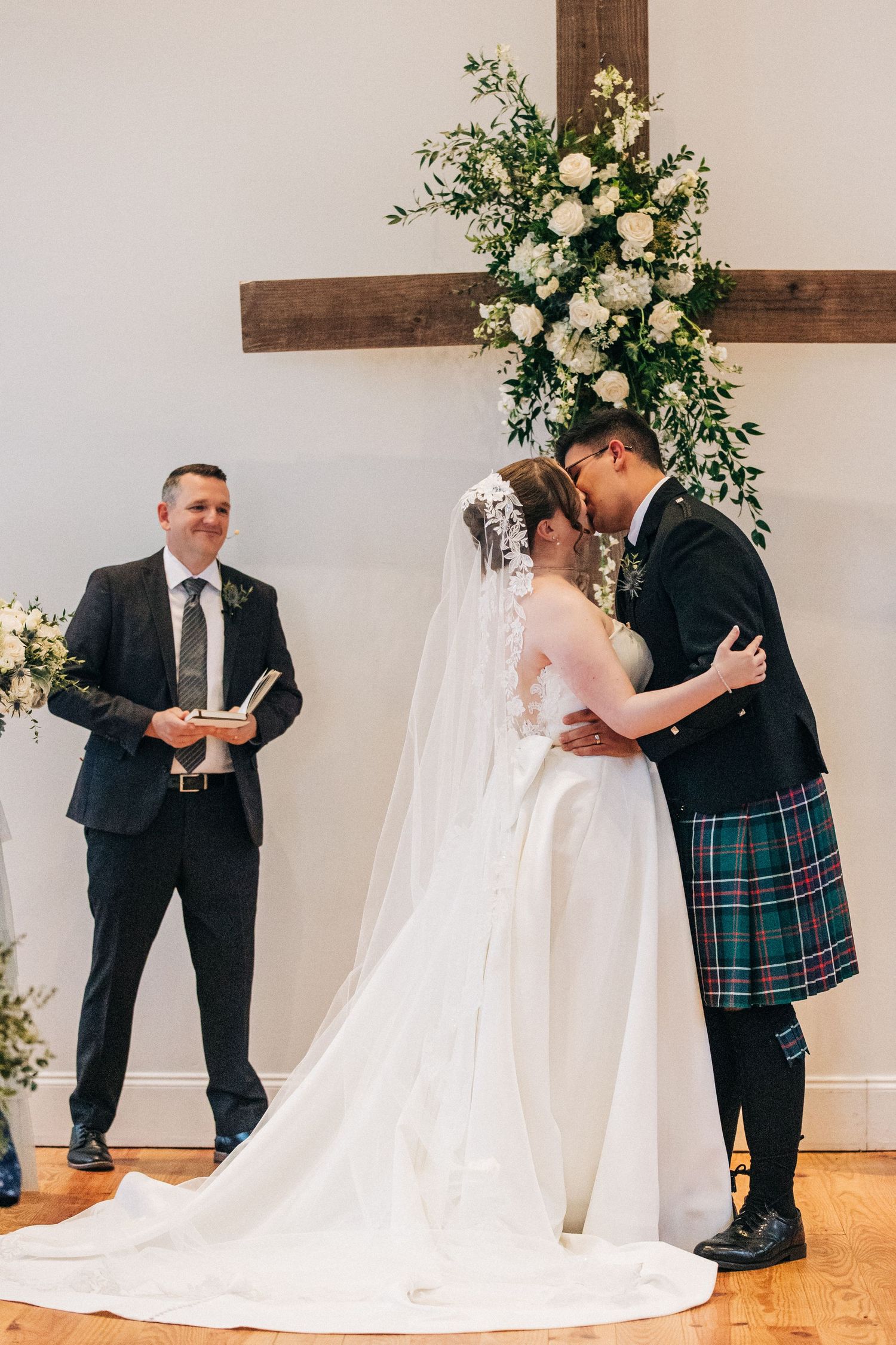 A couple shares their first kiss at the altar during a Scottish-themed wedding ceremony with floral cross backdrop.