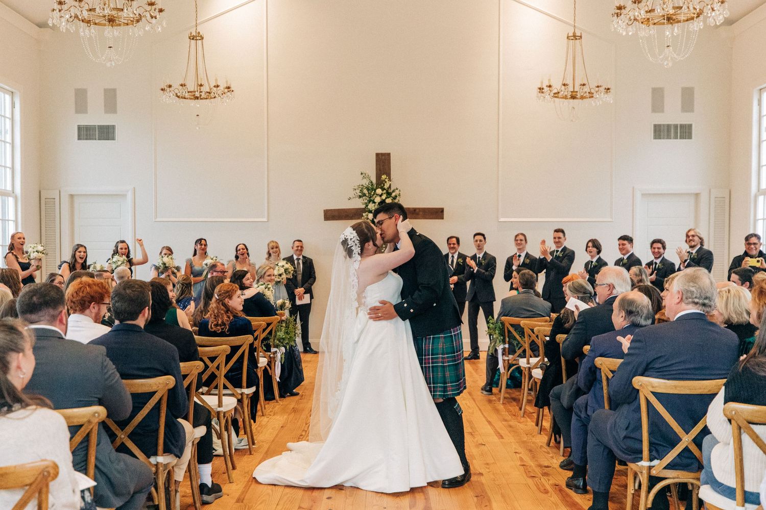 A wedding ceremony in a bright white ballroom with elegant chandeliers and wooden cross-back chairs.