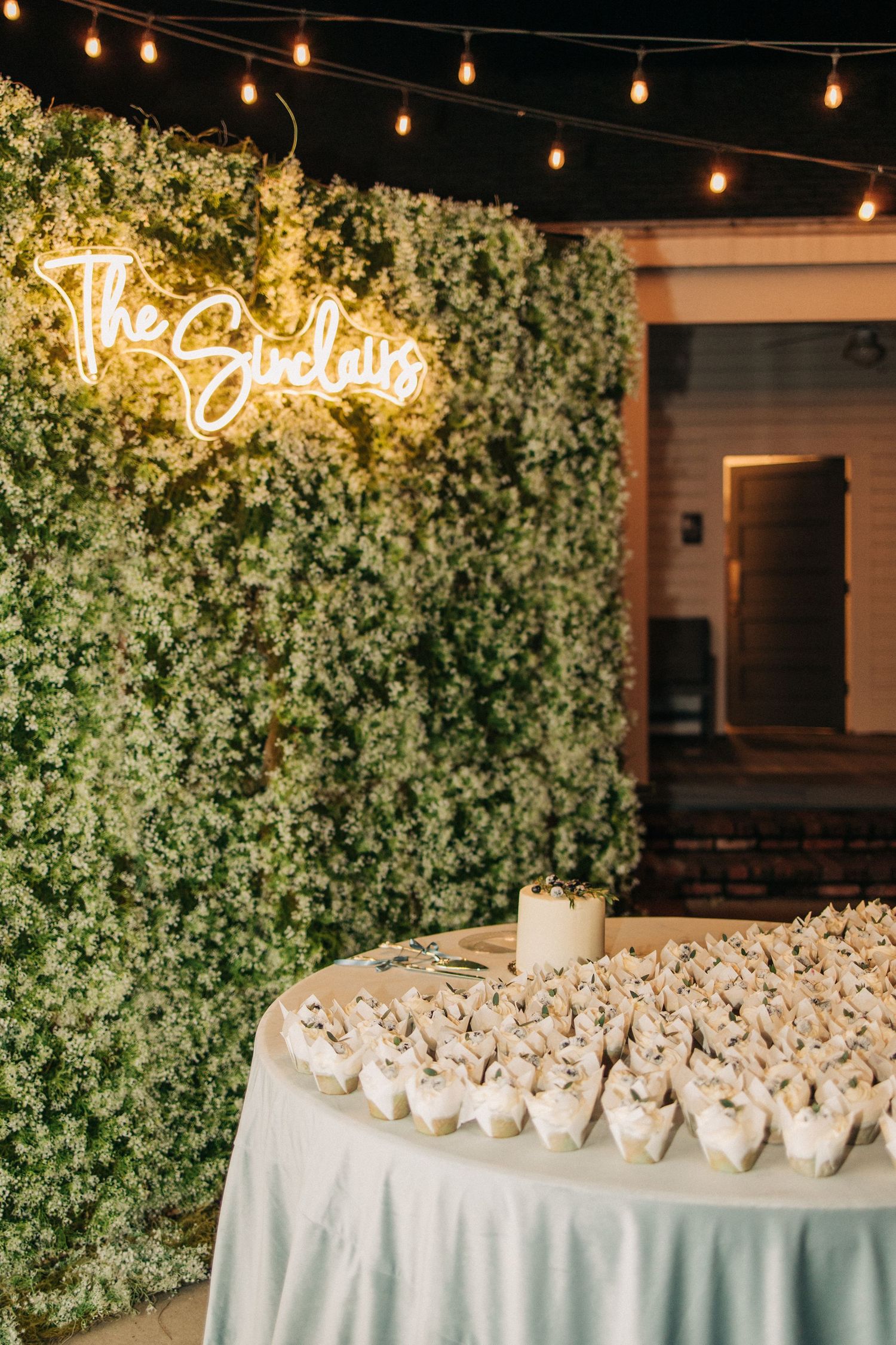 White favor boxes arranged in elegant rows on a table with greenery backdrop and hanging string lights for an event setting.