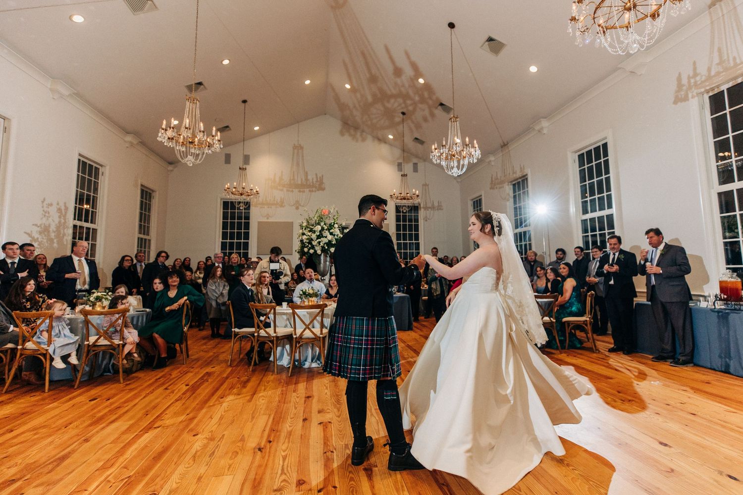 Two people in wedding attire share a dance on a hardwood floor under crystal chandeliers at an elegant reception venue.