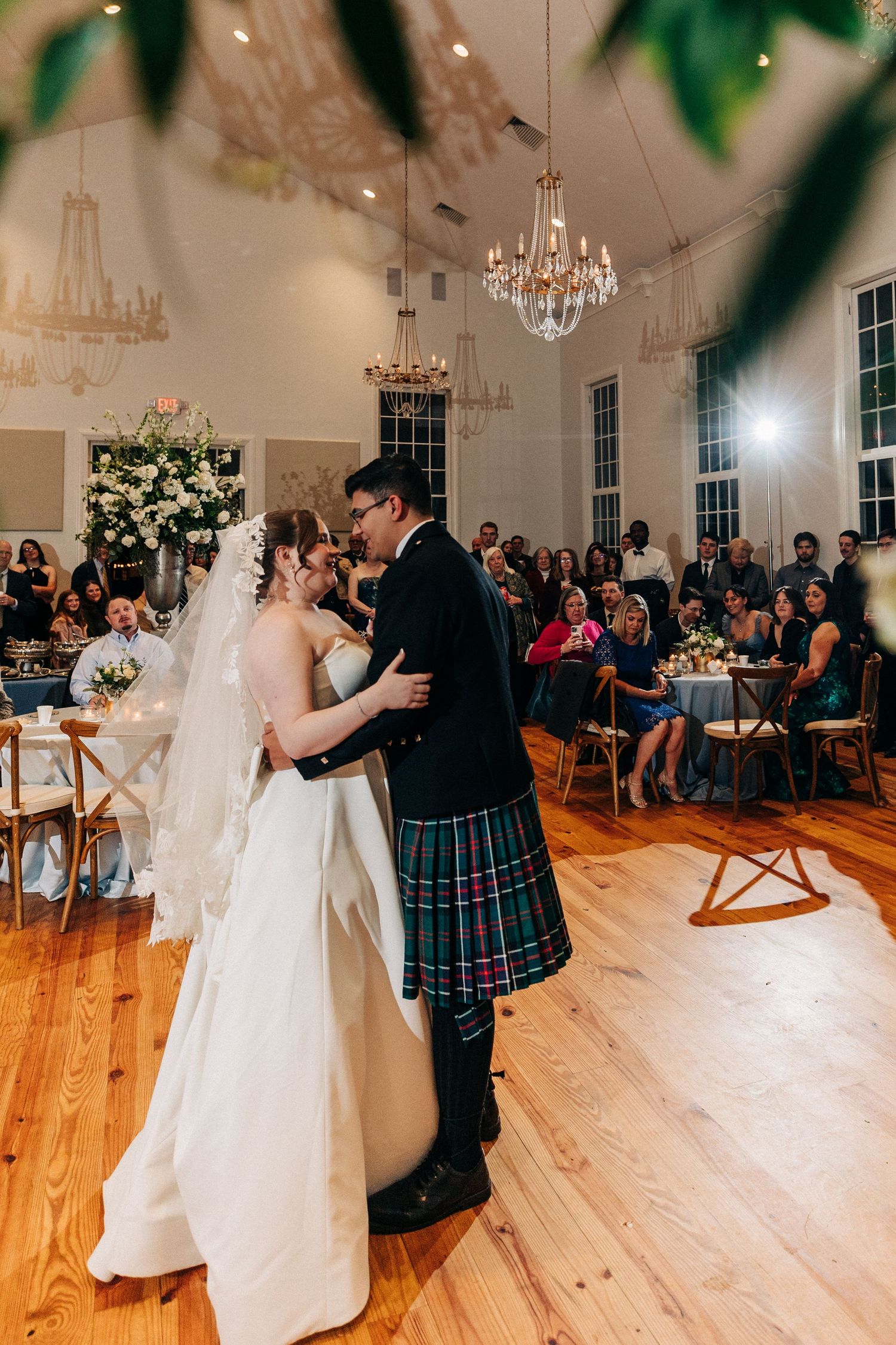 Newlyweds share their first dance in an elegant reception hall with chandeliers and guests seated in wooden chairs.