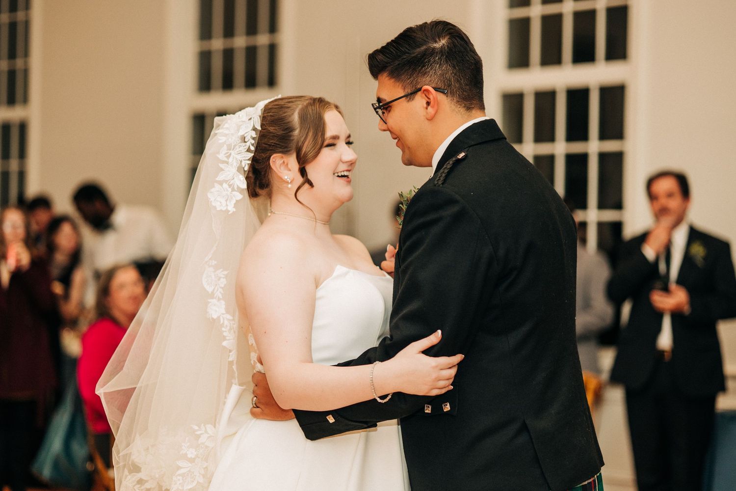 Two wedding guests share a tender dance moment in a ballroom while guests look on in the background.