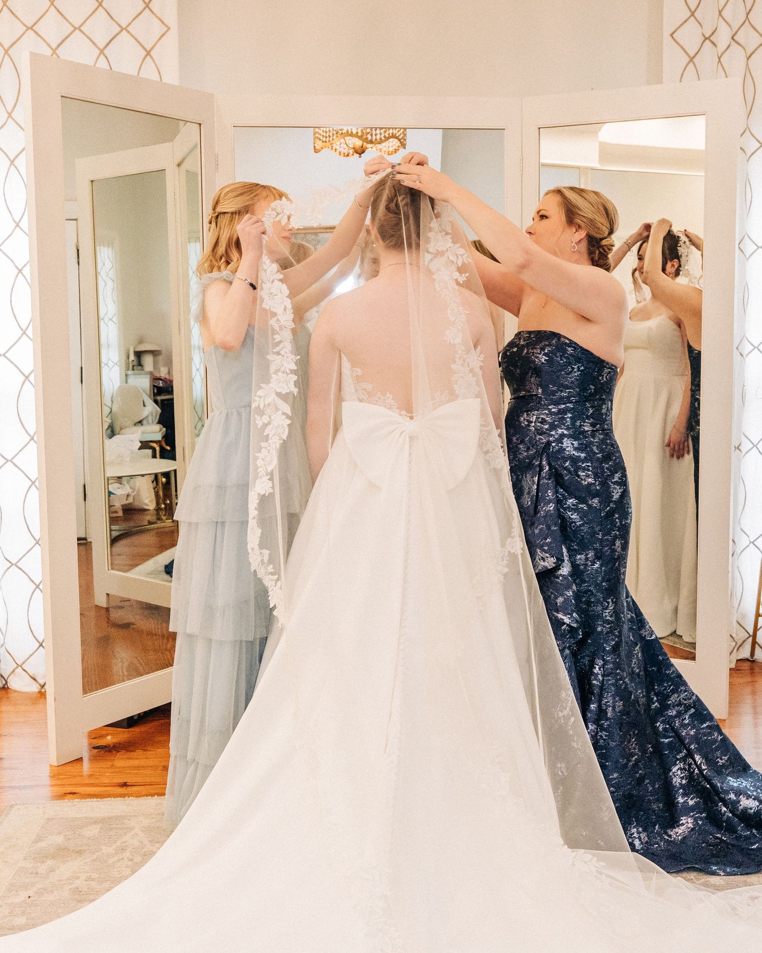 Bridesmaids in grey and navy dresses help adjust the bride's veil in an elegant dressing room with mirrored walls.