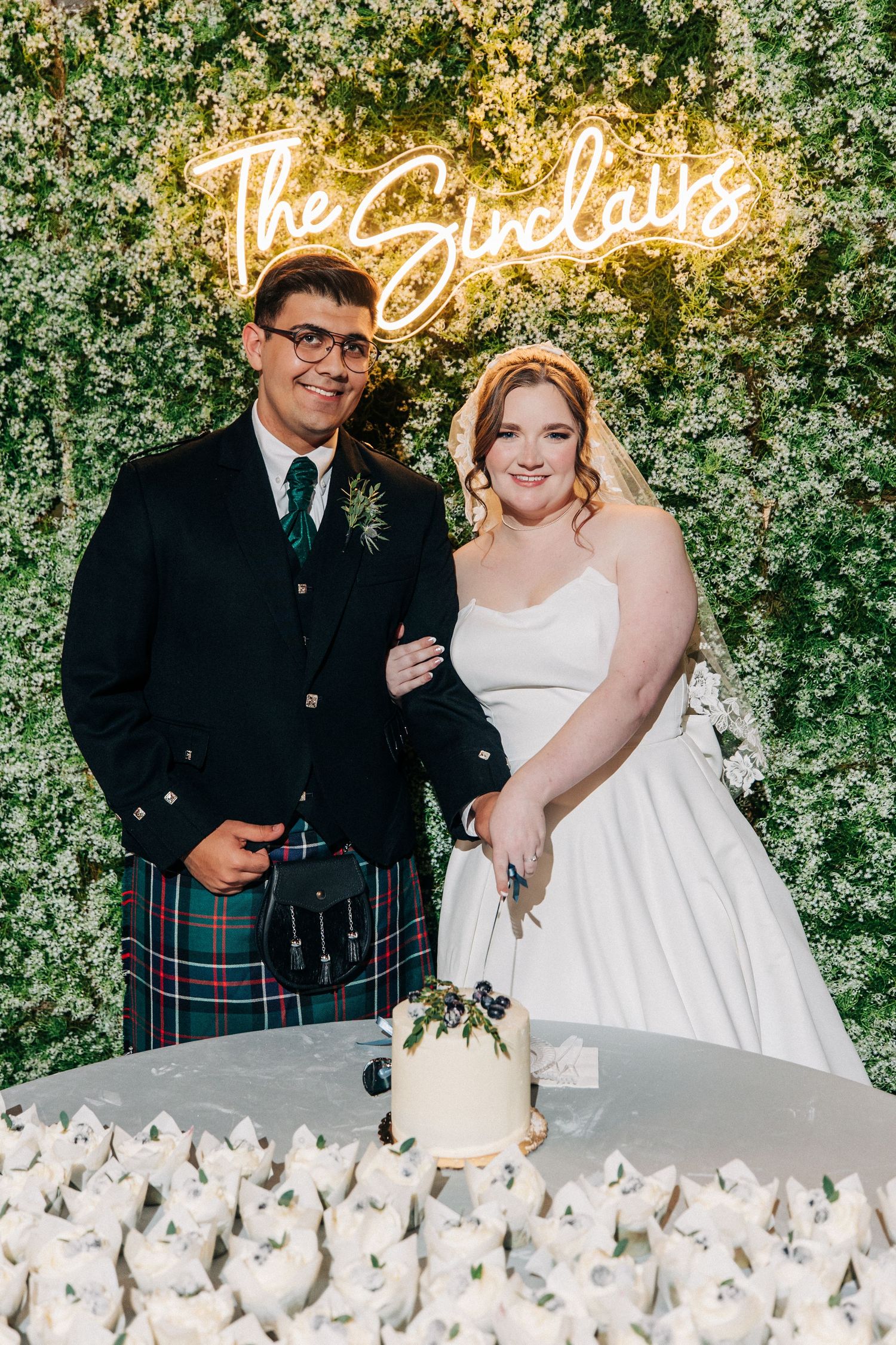 Wedding couple stands by their cake table with a neon sign and greenery wall backdrop as they celebrate their Scottish themed day.